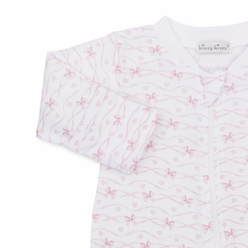 A white long-sleeve baby girl zip-up footed sleeper closeup with a delicate light pink bow and heart pattern and Kissy Kissy brand label.