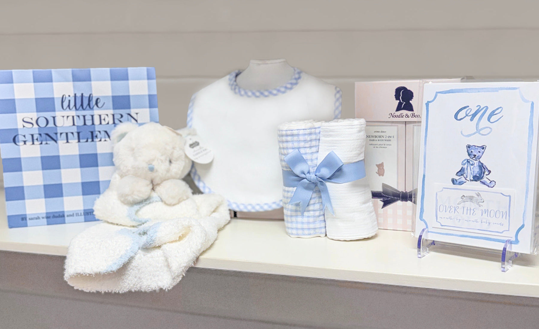 Baby gift set with teddy bear blanket, milestone cards, baby bib, and books on a shelf.