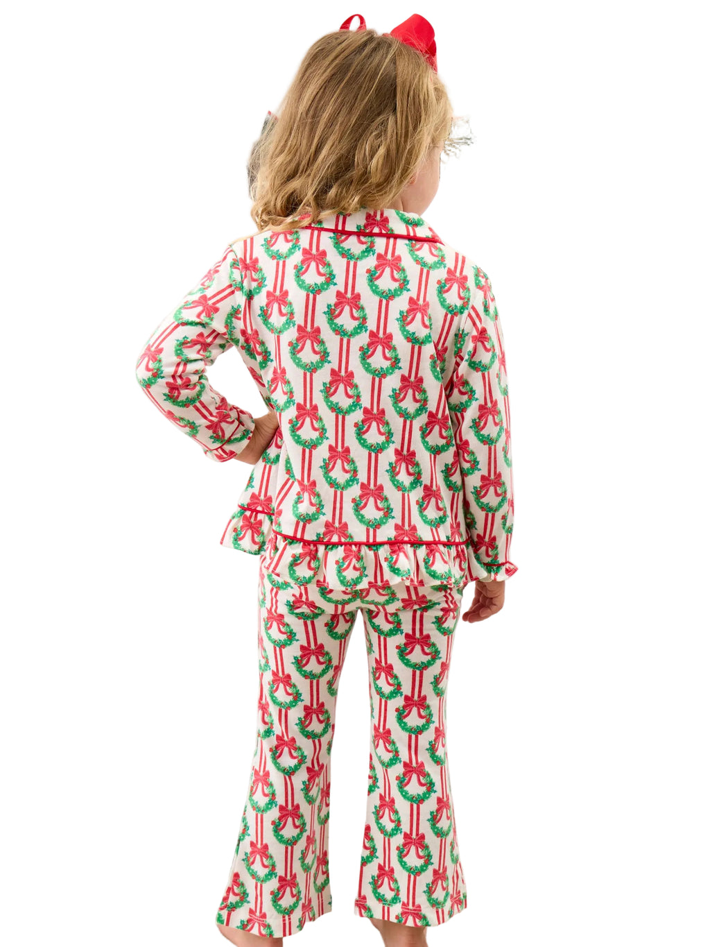 A child standing with their back to the camera, wearing a matching Christmas-themed pajama set with a green wreath and red bow pattern on a white background, in front of a bed with holiday pillows.