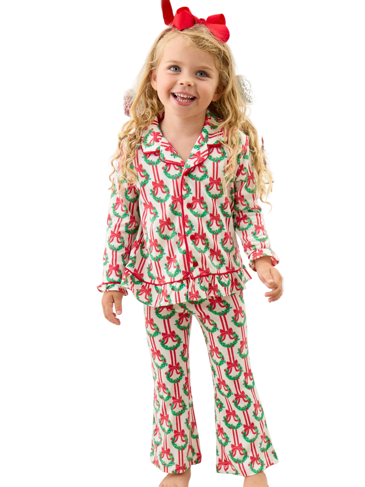 A young girl with long blonde hair and a red bow stands in a bedroom wearing a festive Christmas pajama set with a red and green candy cane and wreath pattern. The background shows a white bed with holiday-themed pillows.