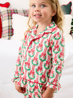 A young girl wearing a festive Christmas-themed pajama set with a red and green wreath pattern, standing in a bedroom setting.
