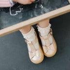Little girl’s shimmery champagne T-strap shoes with bows on top worn with white lace socks