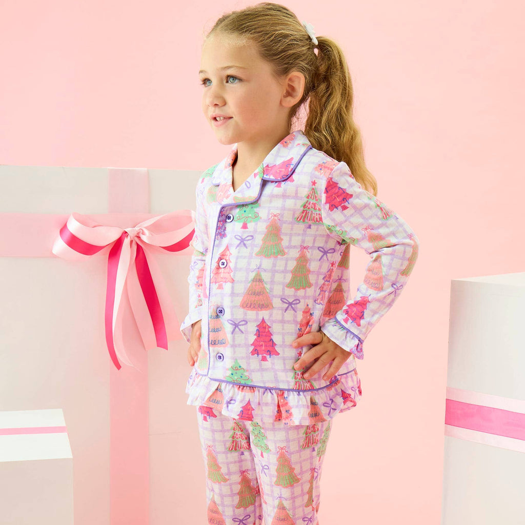 A young girl with blonde hair in a ponytail stands in a pink studio setting wearing a long-sleeve pajama set with a Christmas tree print. The pajamas feature a light pink and white plaid background with colorful Christmas trees and bows.