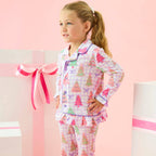 A young girl with blonde hair in a ponytail stands in a pink studio setting wearing a long-sleeve pajama set with a Christmas tree print. The pajamas feature a light pink and white plaid background with colorful Christmas trees and bows.