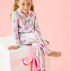 A young girl with blonde pigtails sits on a white pedestal wearing a pink and multicolor dinosaur-patterned pajama set against a solid pink background.