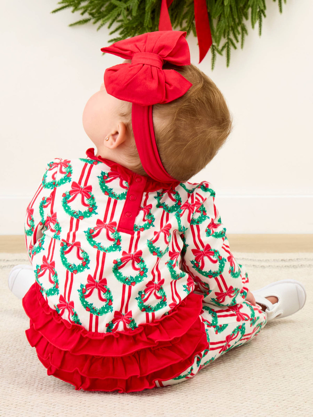 A baby wearing a festive Christmas-themed one-piece outfit with a red bow headband, sitting on a light beige carpet. The outfit features a white base printed with red stripes and green wreaths with red bows, and a solid red ruffled hem.