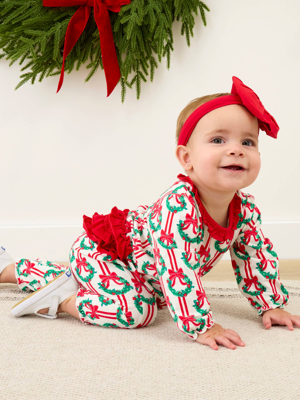 A baby crawling on a light beige carpet wearing a festive Christmas-themed one-piece jumpsuit with a red ruffled bow headband. The jumpsuit has a white background with a green and red holiday wreath and candy cane pattern, and a large red ruffle detail on the back.