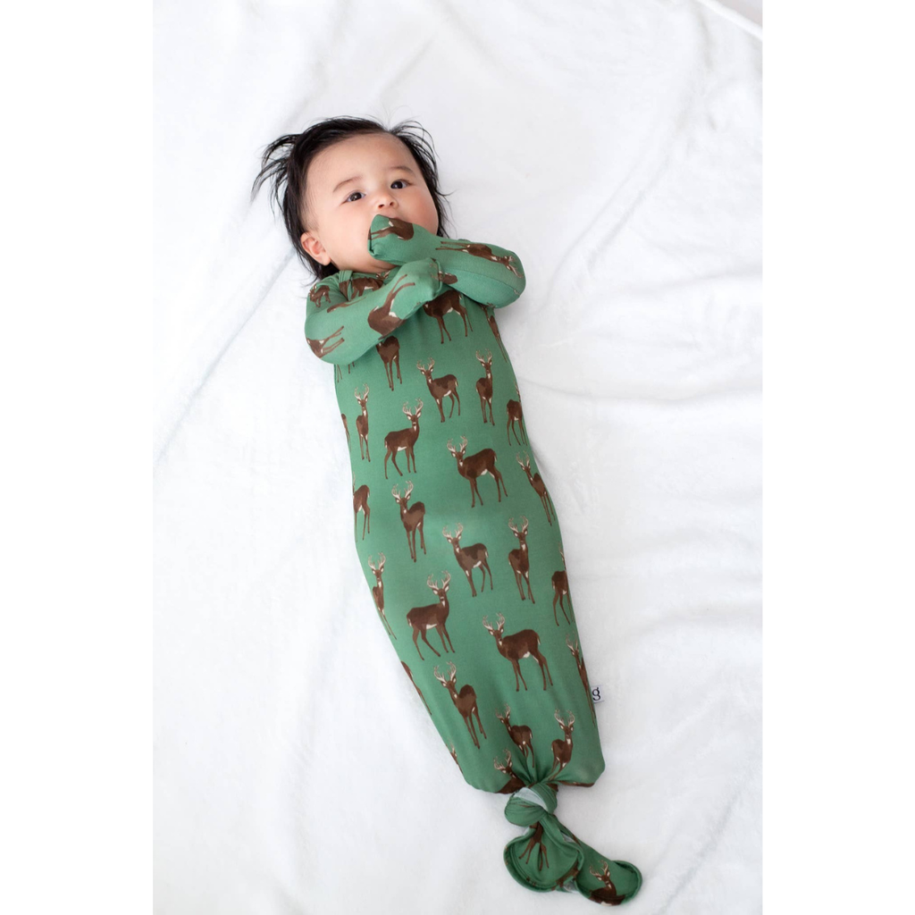 A baby lying on a white blanket, wearing a green footed swaddle blanket with a brown deer pattern.