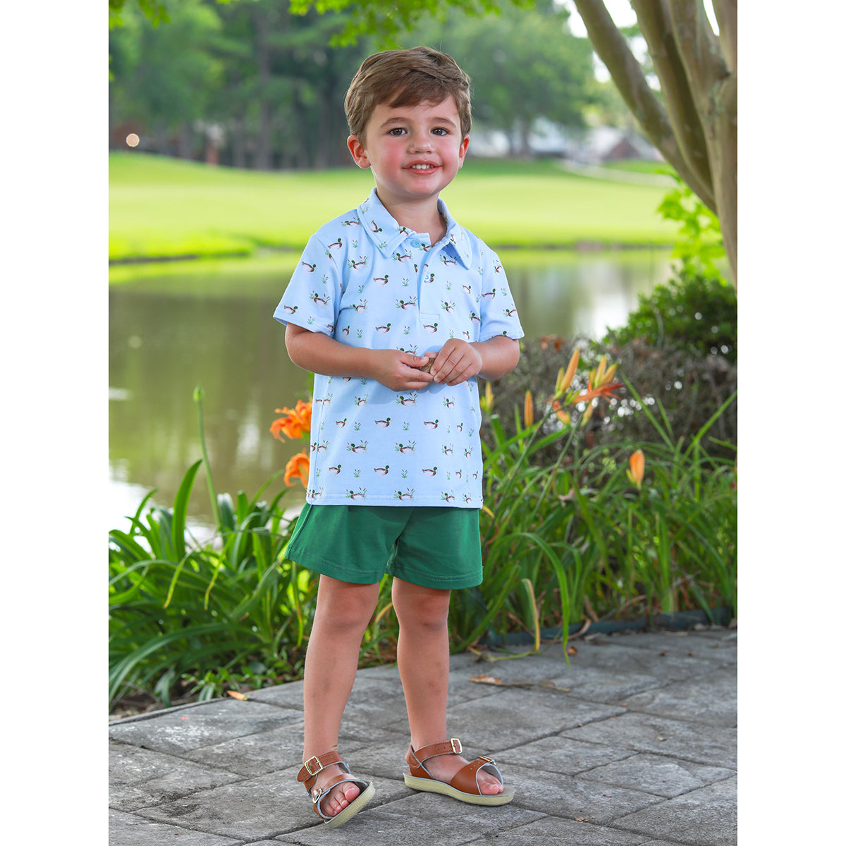 Toddler boy wearing a light blue polo shirt featuring mallard pattern, dark green shorts, and tan sandals.