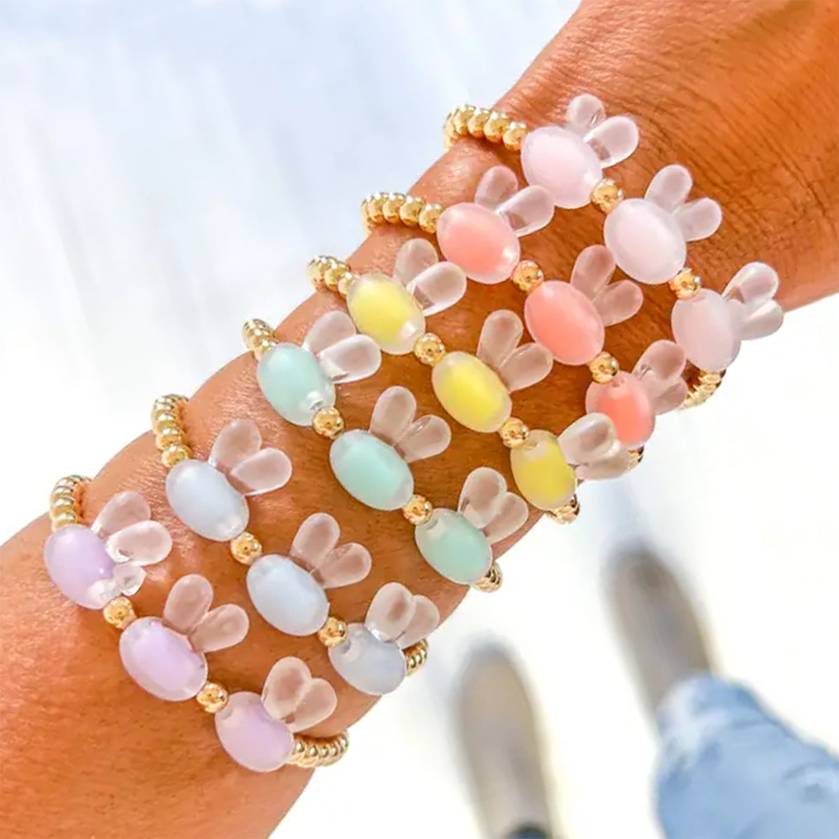 A close-up of a person's wrist wearing multiple pastel beaded stretch bracelets with bunny-ear and oval bead details in pink, yellow, mint, and lavender, separated by small gold spacer beads.