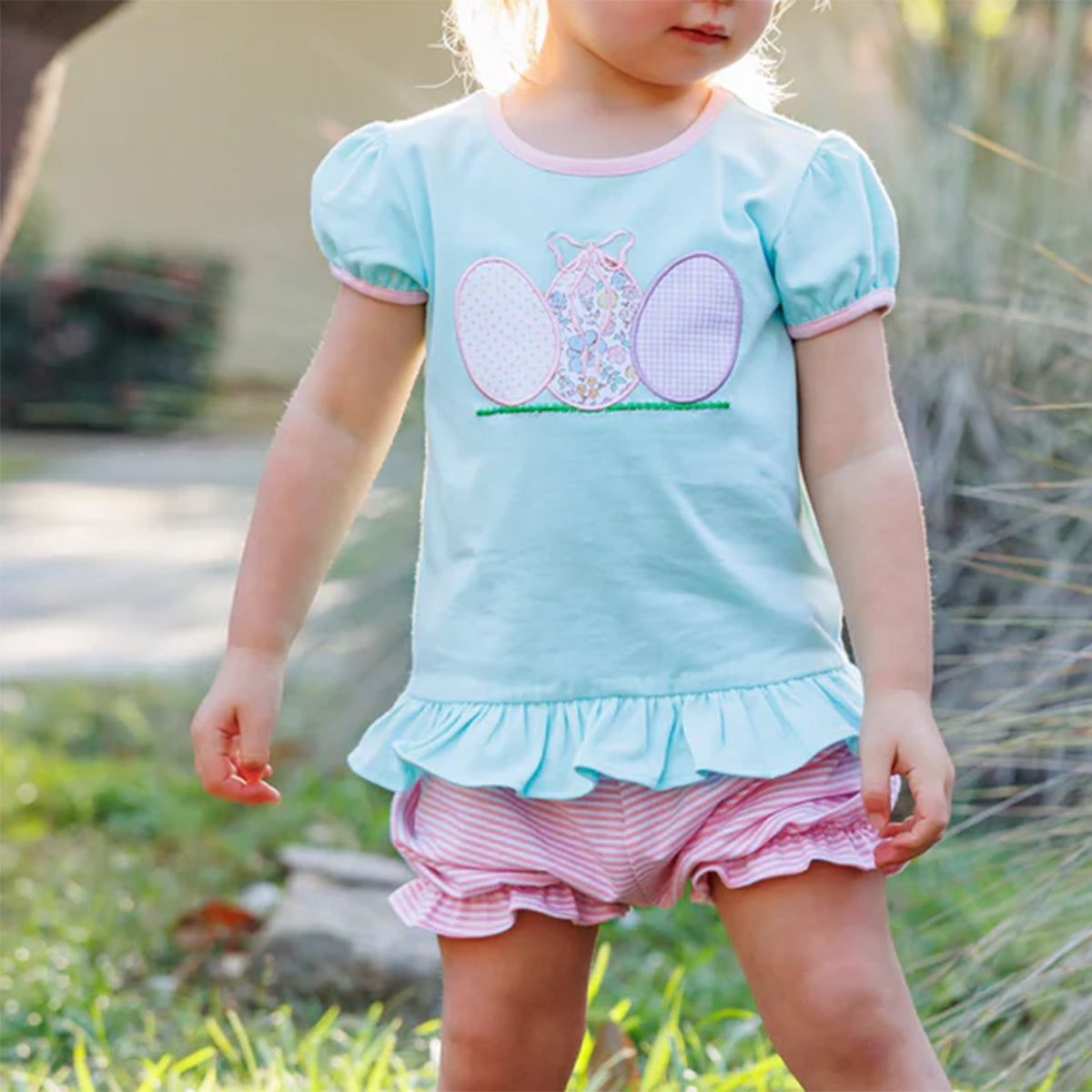 Child wearing a Mint green short sleeve shirt with ruffles, pink trim and appliqued Easter eggs with pink and white stripe bloomers.