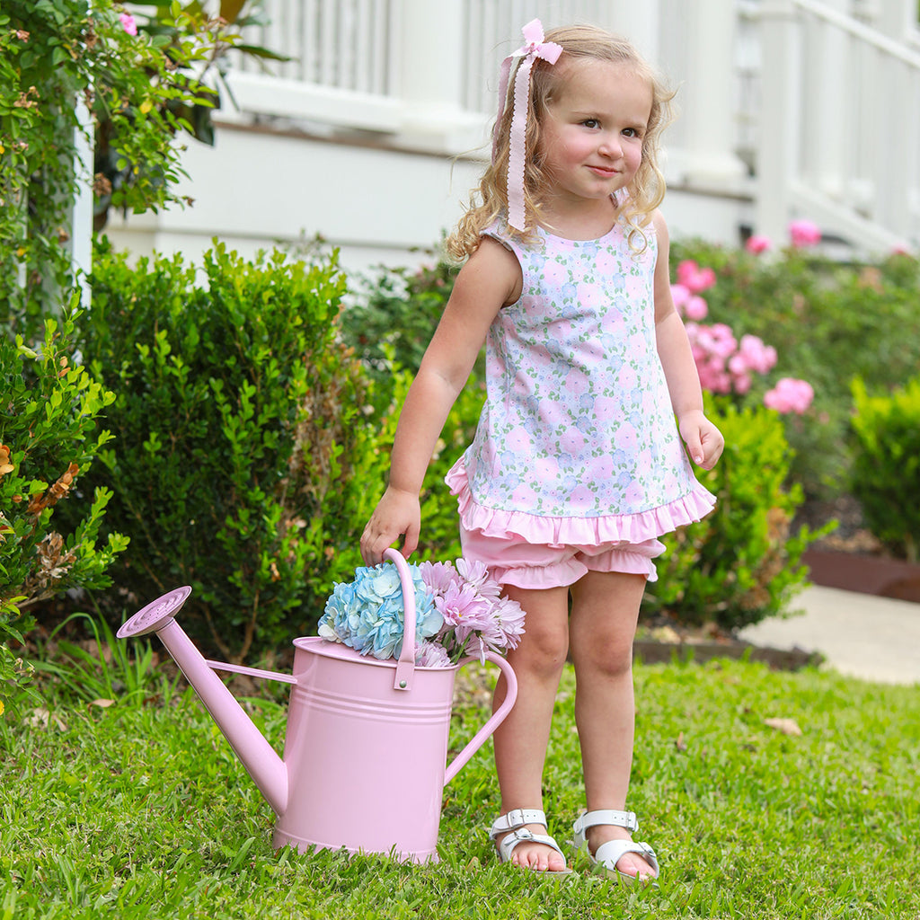 Little girl wearing a floral knit bloomer set carrying a pink watering can with hydrangeas.
