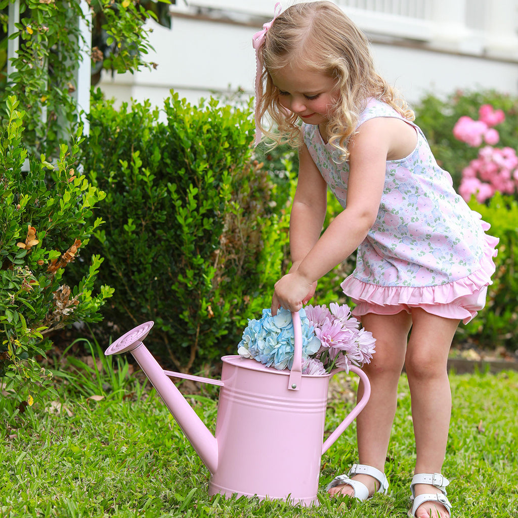 Little girl wearing a floral knit bloomer set carrying a pink watering can with hydrangeas.