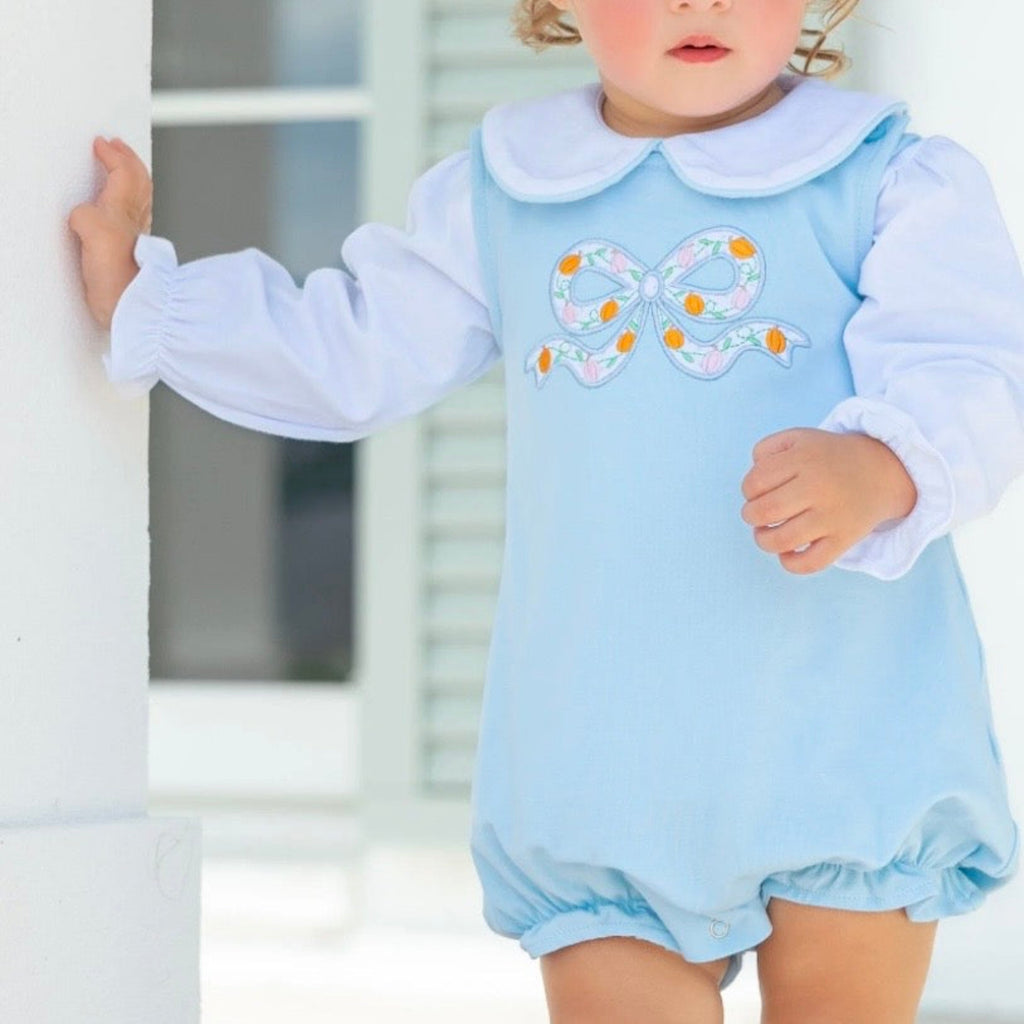 A toddler girl wearing a light blue bubble with a pumpkin bow applique on the chest and a long sleeve white knit shirt with Peter Pan collar.