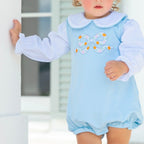 A toddler girl wearing a light blue bubble with a pumpkin bow applique on the chest and a long sleeve white knit shirt with Peter Pan collar.