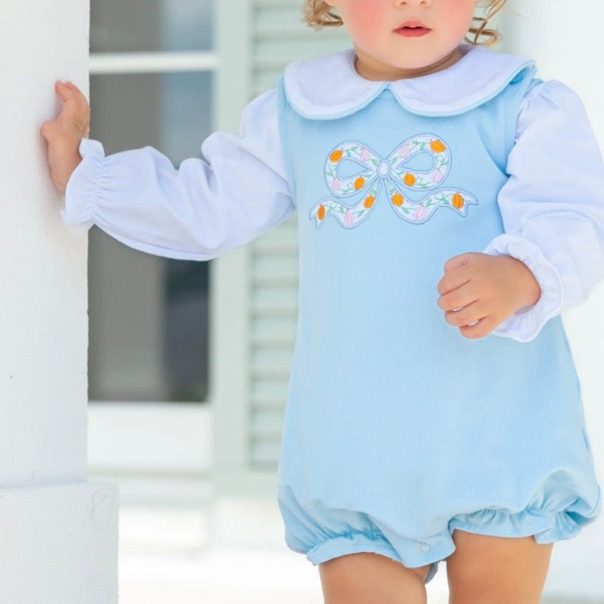 A toddler girl wearing a light blue bubble with a pumpkin bow applique on the chest and a long sleeve white knit shirt with Peter Pan collar.
