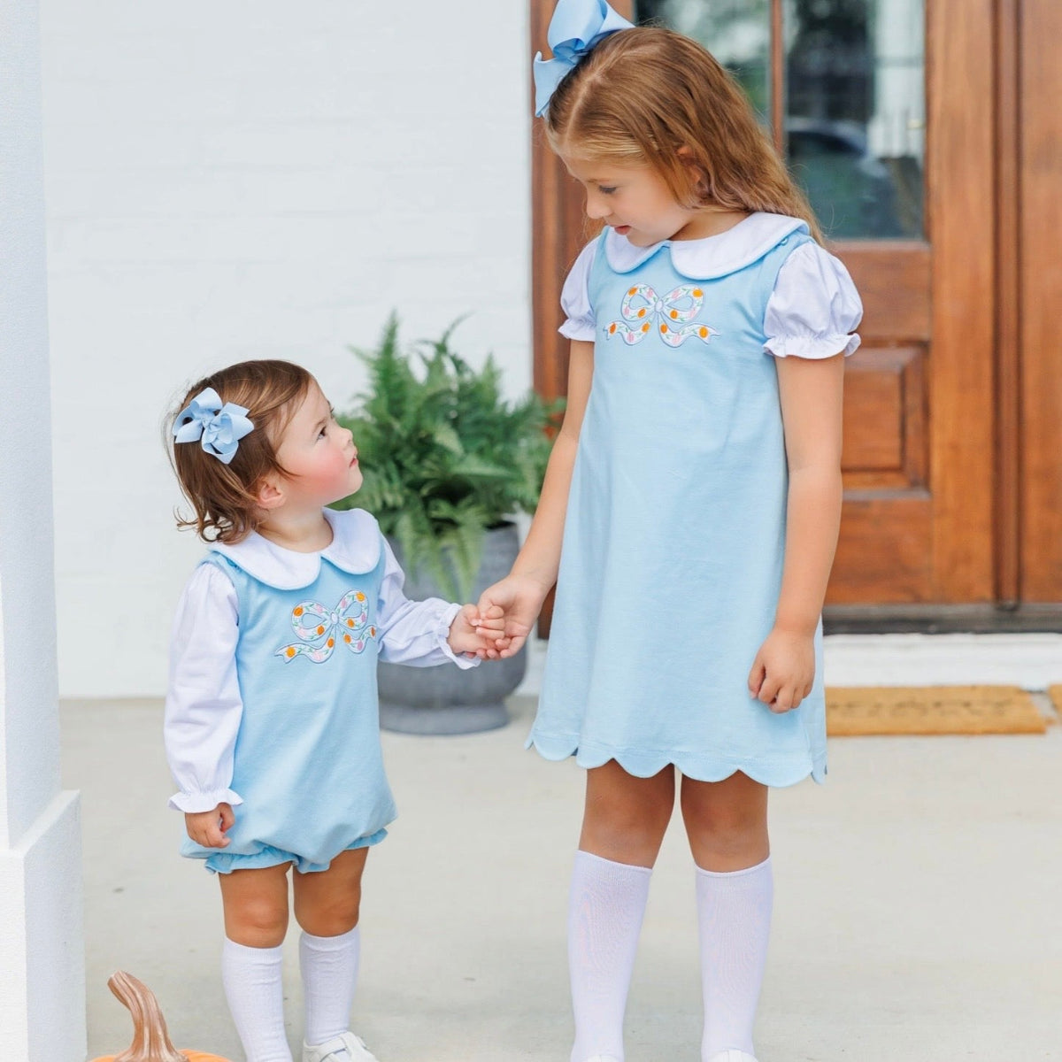 Two little girls wearing matching light blue and white fall outfits that feature an appliqued bow with embroideredd pumpkins.