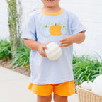 A little boy wearing a light blue and white striped t-shirt with a pumpkin graphic and light orange shorts.