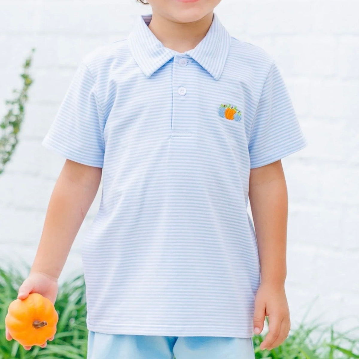 A little boy wearing a light blue and white striped short-sleeve polo shirt featuring embroidered pumpkins on the left chest.