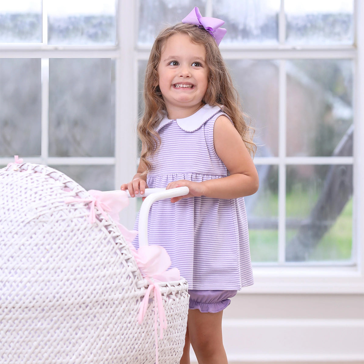 Toddler girl wearing a sleeveless lavender stripe knit bloomer set with a white collar pushing a baby carriage.