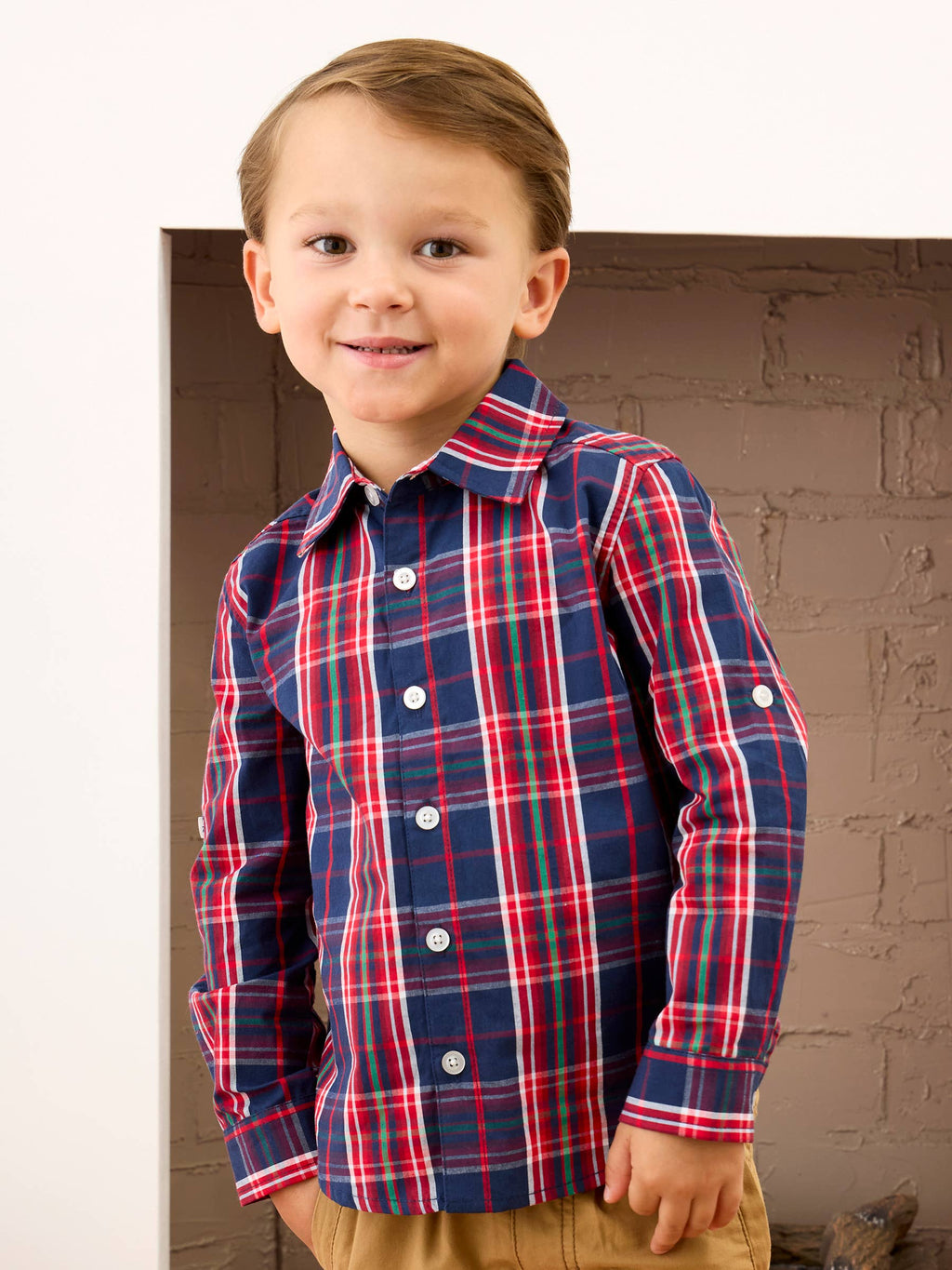 A young boy wearing a long-sleeve plaid button-down shirt in navy blue with red, white, and green stripes, paired with tan pants, standing in front of a brick fireplace.