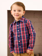 A young boy wearing a long-sleeve plaid button-down shirt in navy blue with red, white, and green stripes, paired with tan pants, standing in front of a brick fireplace.