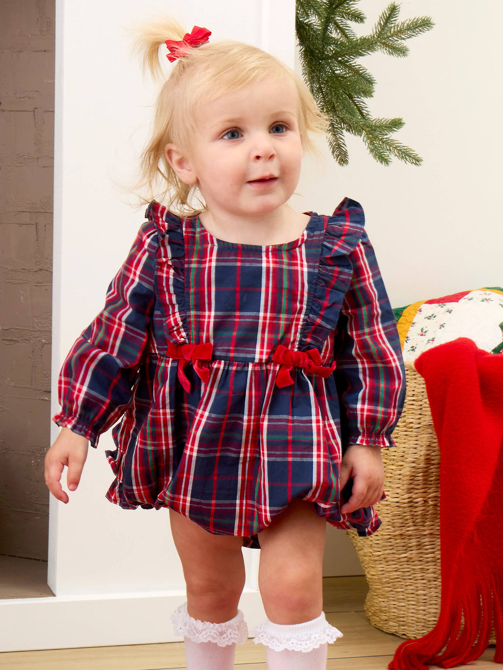 A toddler girl wearing a red, navy, and white plaid long-sleeve romper dress with ruffled shoulders and red bow details at the waist, standing in an indoor holiday setting.