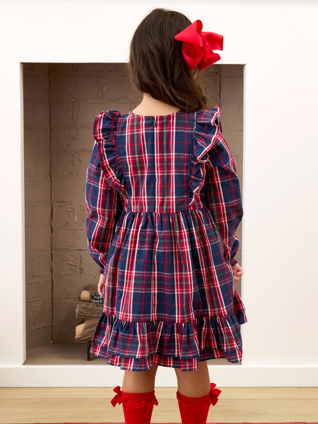 A young girl viewed from behind wearing a navy blue plaid dress with red and white stripes, featuring ruffled shoulders and a tiered skirt. She has a large red bow in her hair and is wearing red socks, standing in front of a white wall with a brick fireplace.