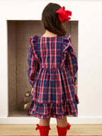 A young girl viewed from behind wearing a navy blue plaid dress with red and white stripes, featuring ruffled shoulders and a tiered skirt. She has a large red bow in her hair and is wearing red socks, standing in front of a white wall with a brick fireplace.