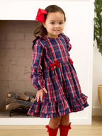 A young girl wearing a red and navy plaid long-sleeve dress with ruffled shoulders and waist, red bow accents, and red knee-high socks, standing in front of a fireplace.