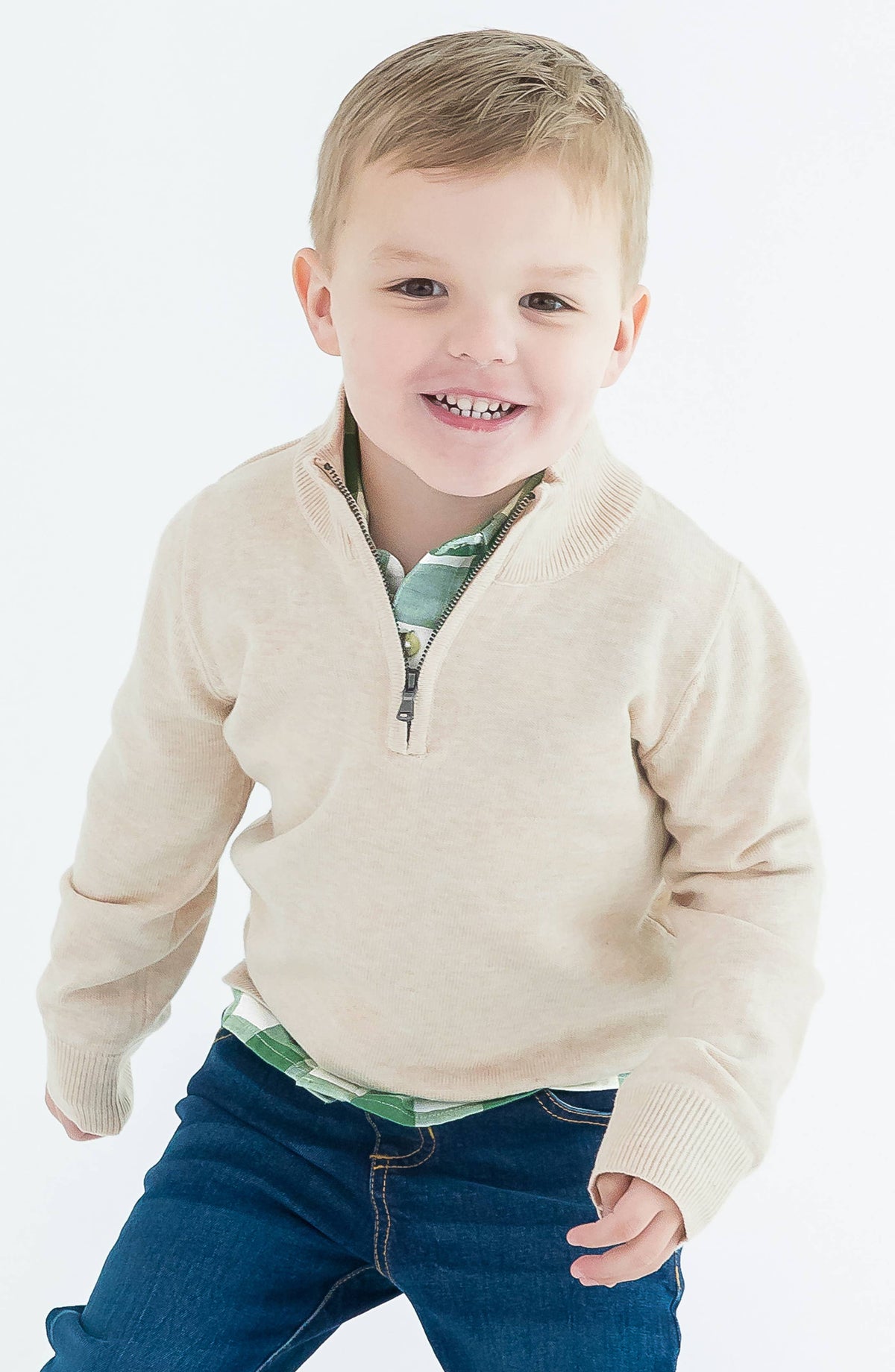 Child wearing a beige sweater over a green shirt with blue jeans on a white background
