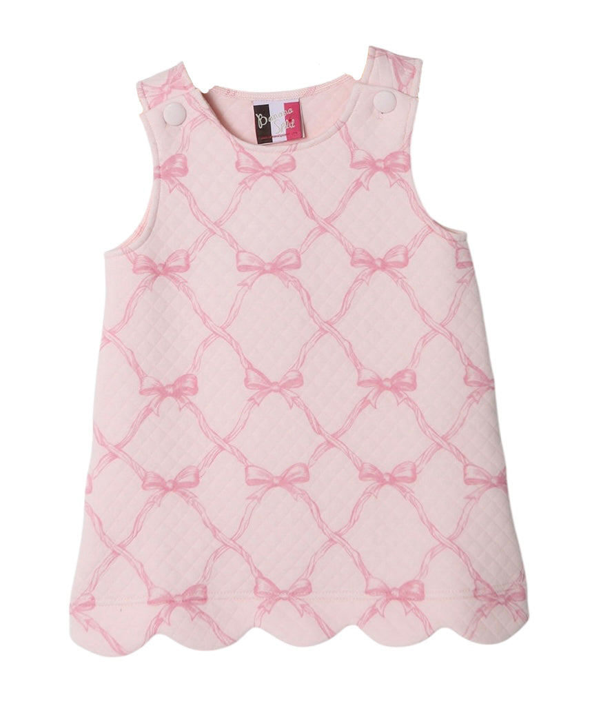 A light pink sleeveless girls jumper dress with a scalloped hem, featuring a pink trellis pattern with bow motifs and buttons on the shoulders.