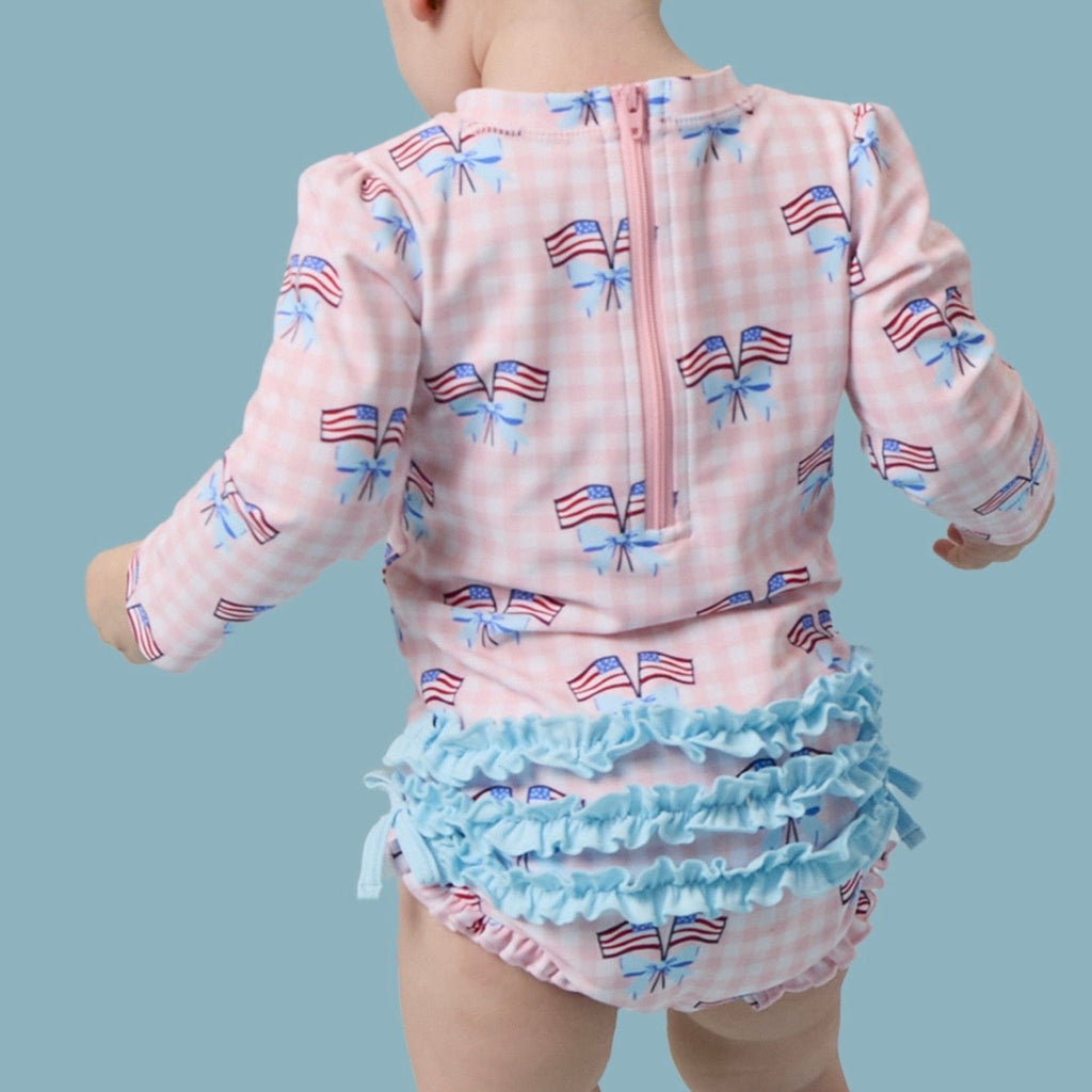 Child wearing a long sleeve bathing suit with American flag patterns on a pink gingham check background.