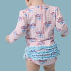 Child wearing a long sleeve bathing suit with American flag patterns on a pink gingham check background.