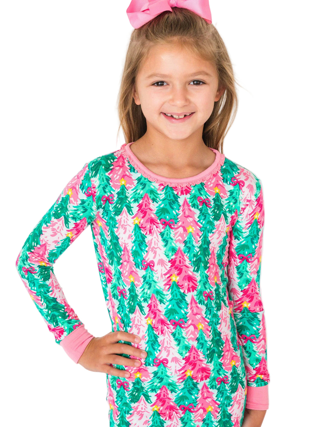 A young girl with a pink bow in her hair is wearing a long-sleeve pajama top featuring a colorful pine tree pattern in green, pink, and white, set against a plain white background.