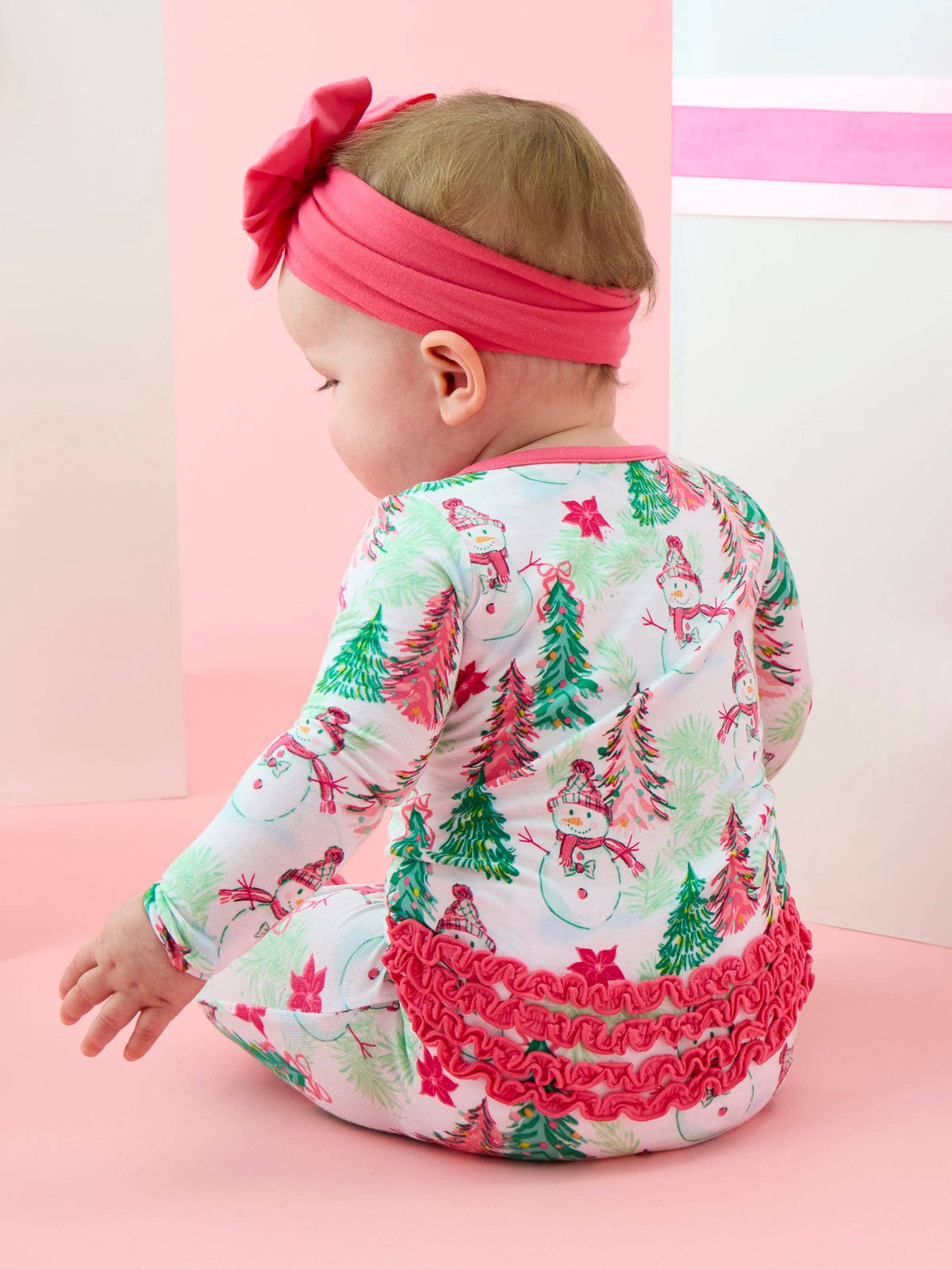 A baby wearing a festive Christmas-themed long-sleeve romper and a matching pink bow headband, sitting on a pink studio floor.