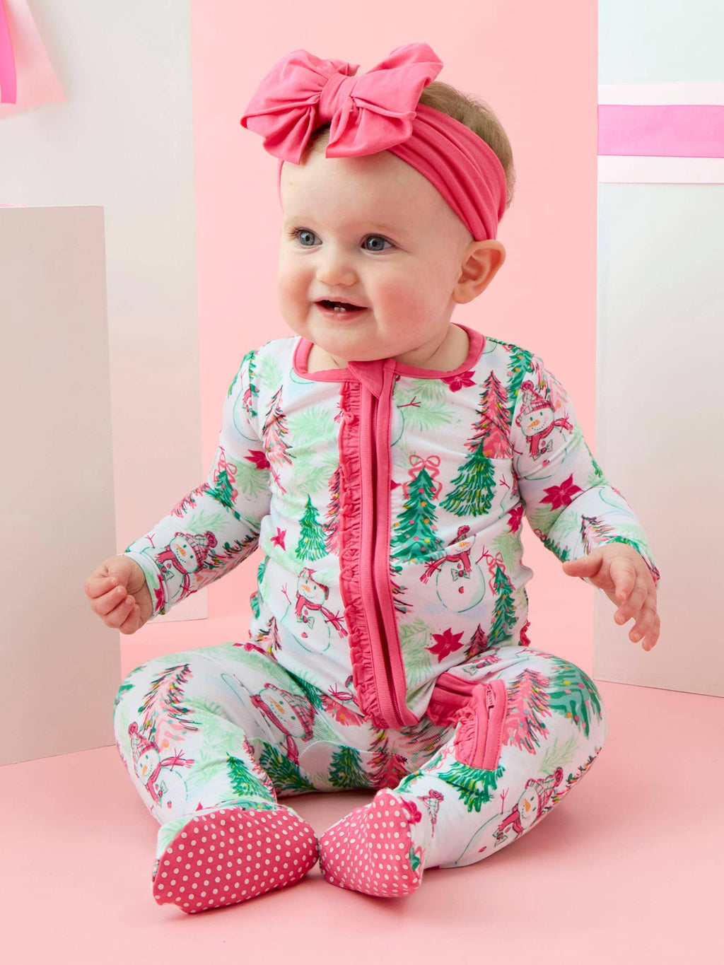 A baby wearing a festive Christmas-themed footed sleeper with a pink zipper and ruffle trim, matching pink polka-dot booties, and a pink bow headband, sitting against a pastel pink and white background.