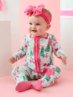A baby wearing a festive Christmas-themed footed sleeper with a pink zipper and ruffle trim, matching pink polka-dot booties, and a pink bow headband, sitting against a pastel pink and white background.