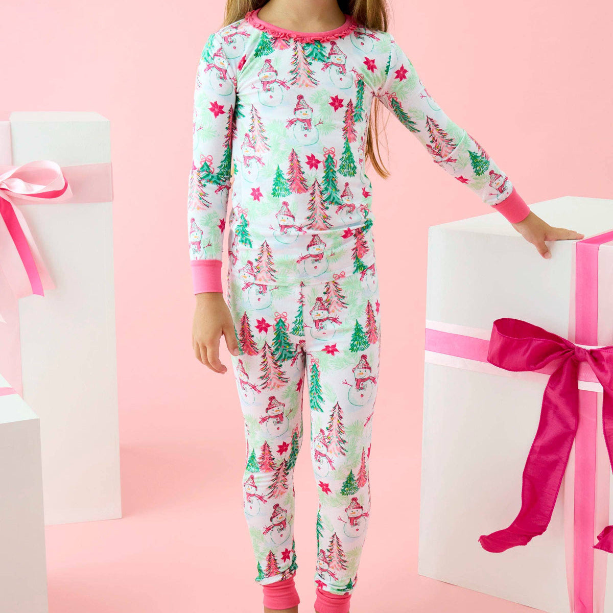 A young girl with a pink bow in her hair stands in a studio setting wearing a long-sleeve Christmas-themed pajama set. The pajamas are white with a pattern of green and pink Christmas trees and stars, with pink trim on the collar, cuffs, and ankles.