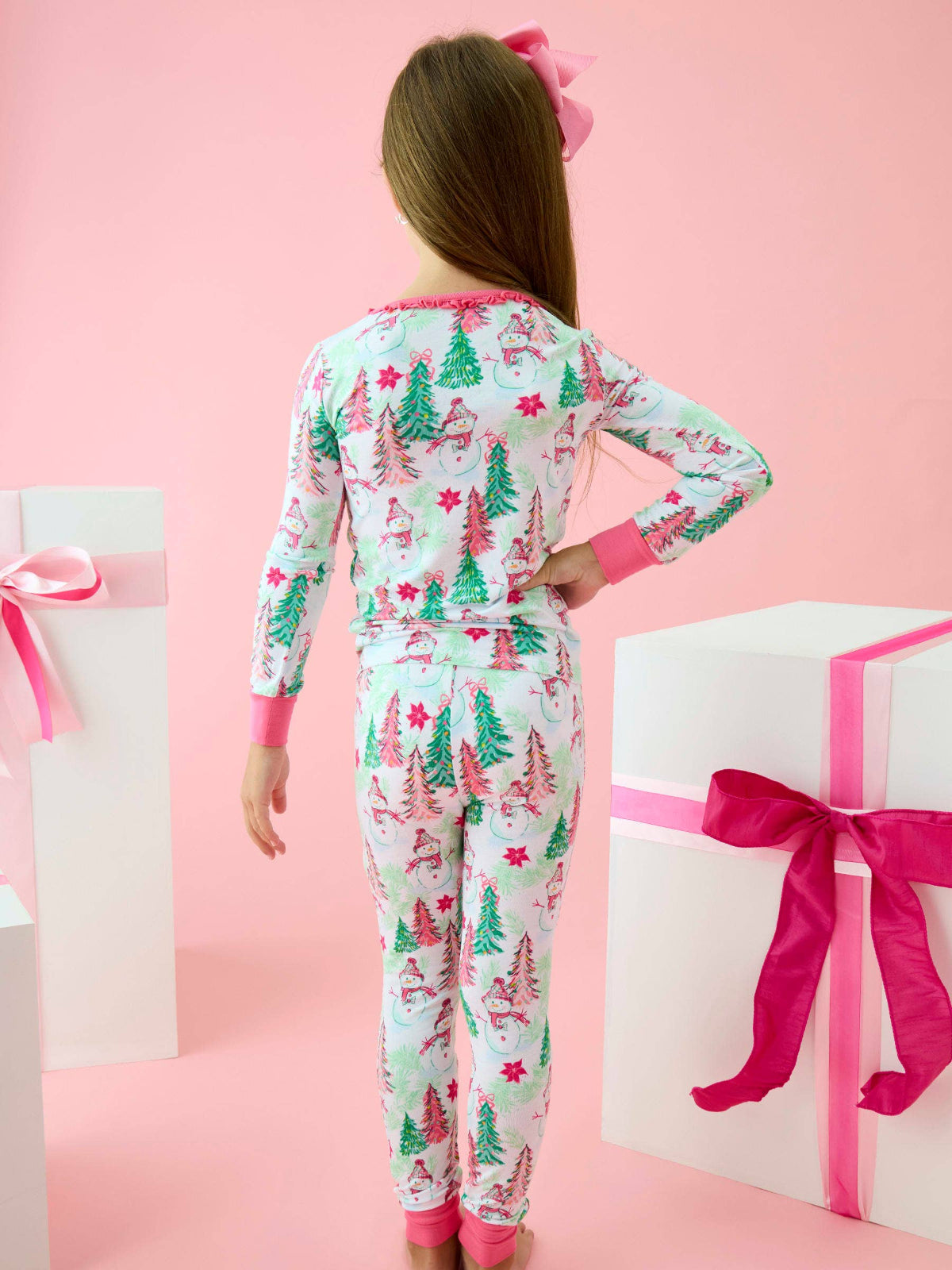 A child wearing a long-sleeve pajama set with a Christmas tree and snowman print, shown from the back against a pink background with gift boxes.