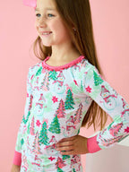 A young girl wearing a long-sleeve white pajama top with a colorful Christmas print of snowmen, pine trees, and stars, posed against a solid pink background.