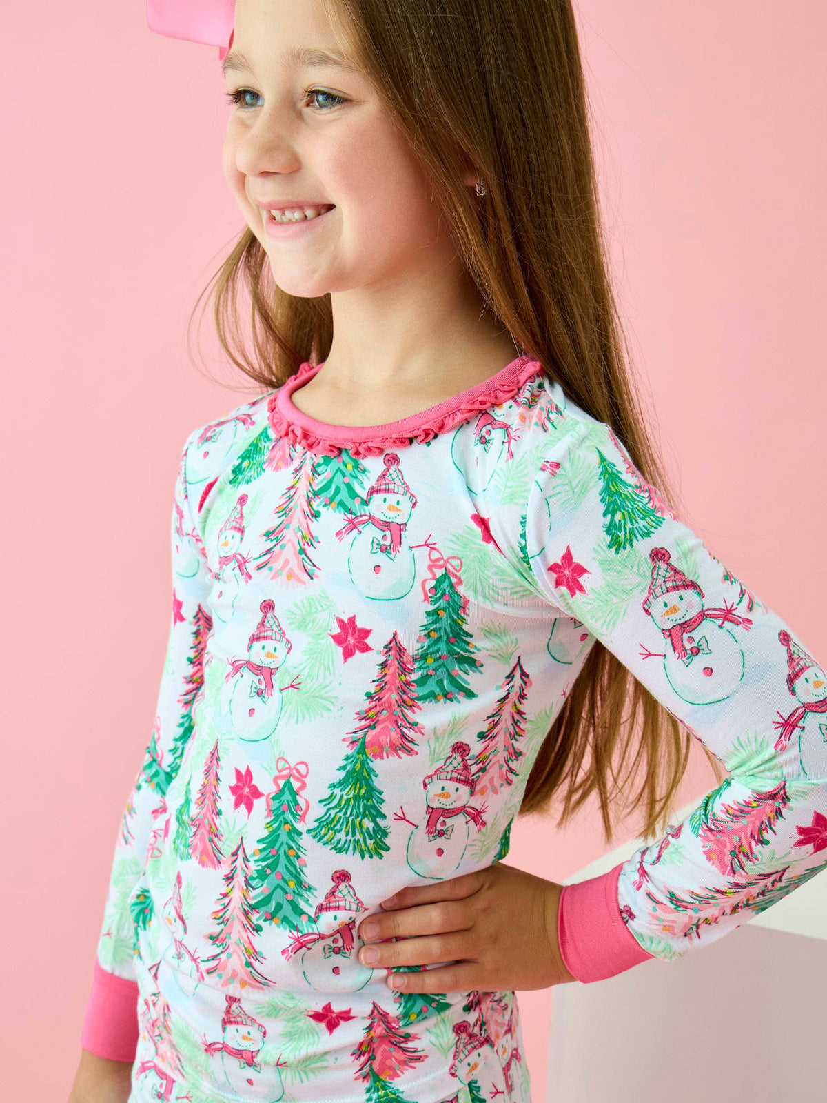 A young girl wearing a long-sleeve white pajama top with a colorful Christmas print of snowmen, pine trees, and stars, posed against a solid pink background.