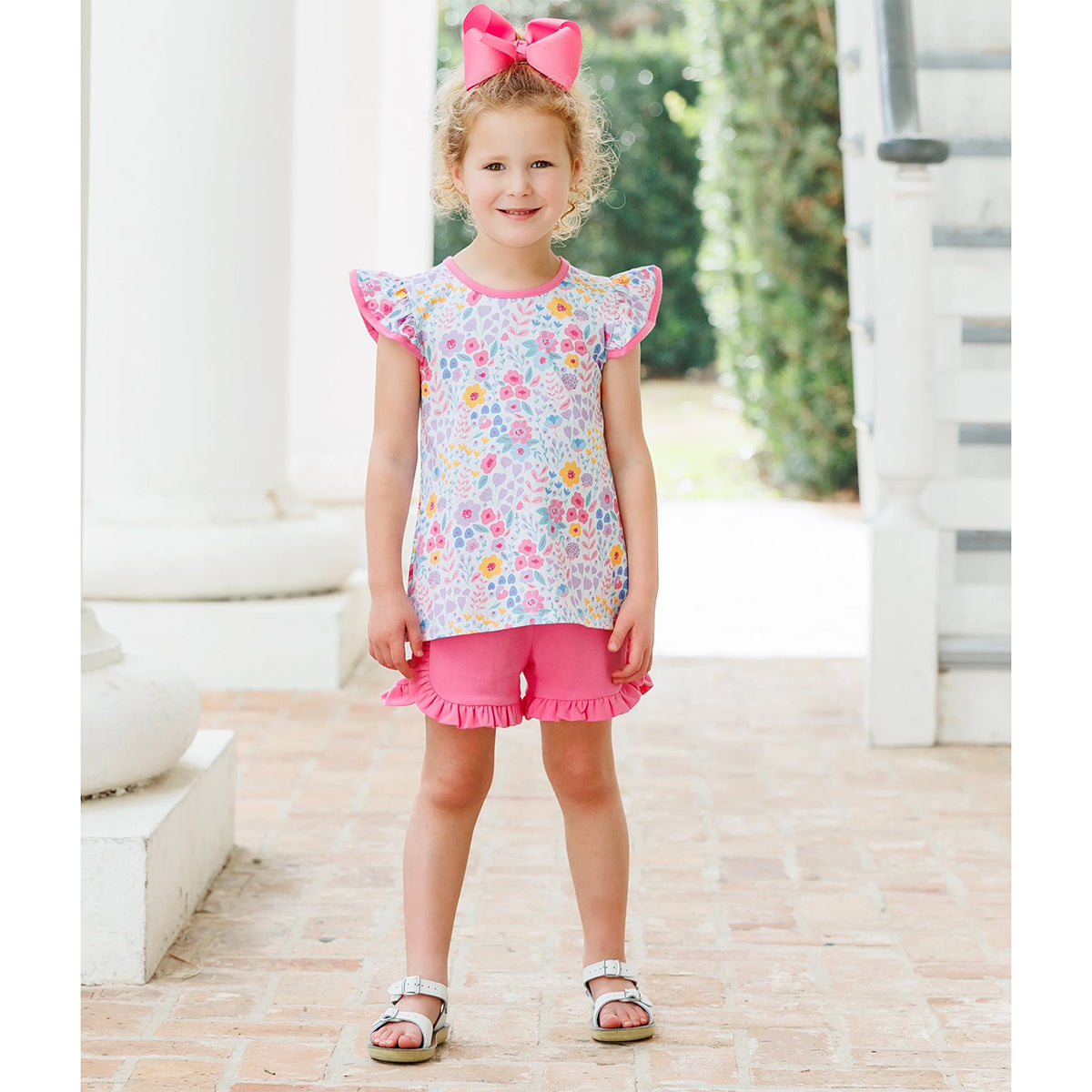 Little girl wearing a floral top and ruffled pink shorts set, white sandals, and a bright pink hairbow.