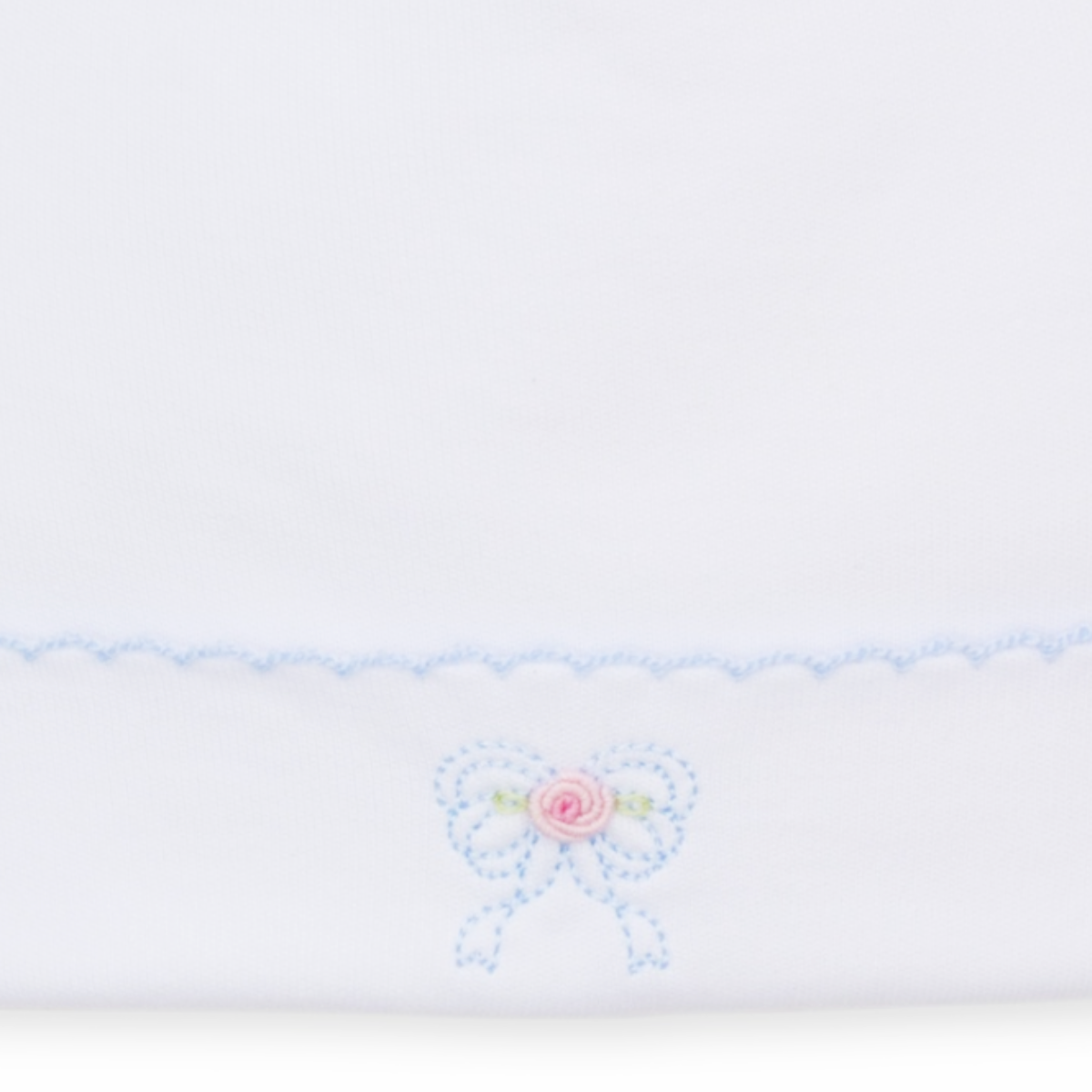 A white knit newborn baby girl hat closeup featuring a light blue scalloped trim and a small embroidered bow with a pink rose center.