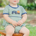 A baby boy wearing a fall bubble in green and white stripes, featuring a steel blue crew neck and cuffs and an appliqued tractor and orange pumpkin design.