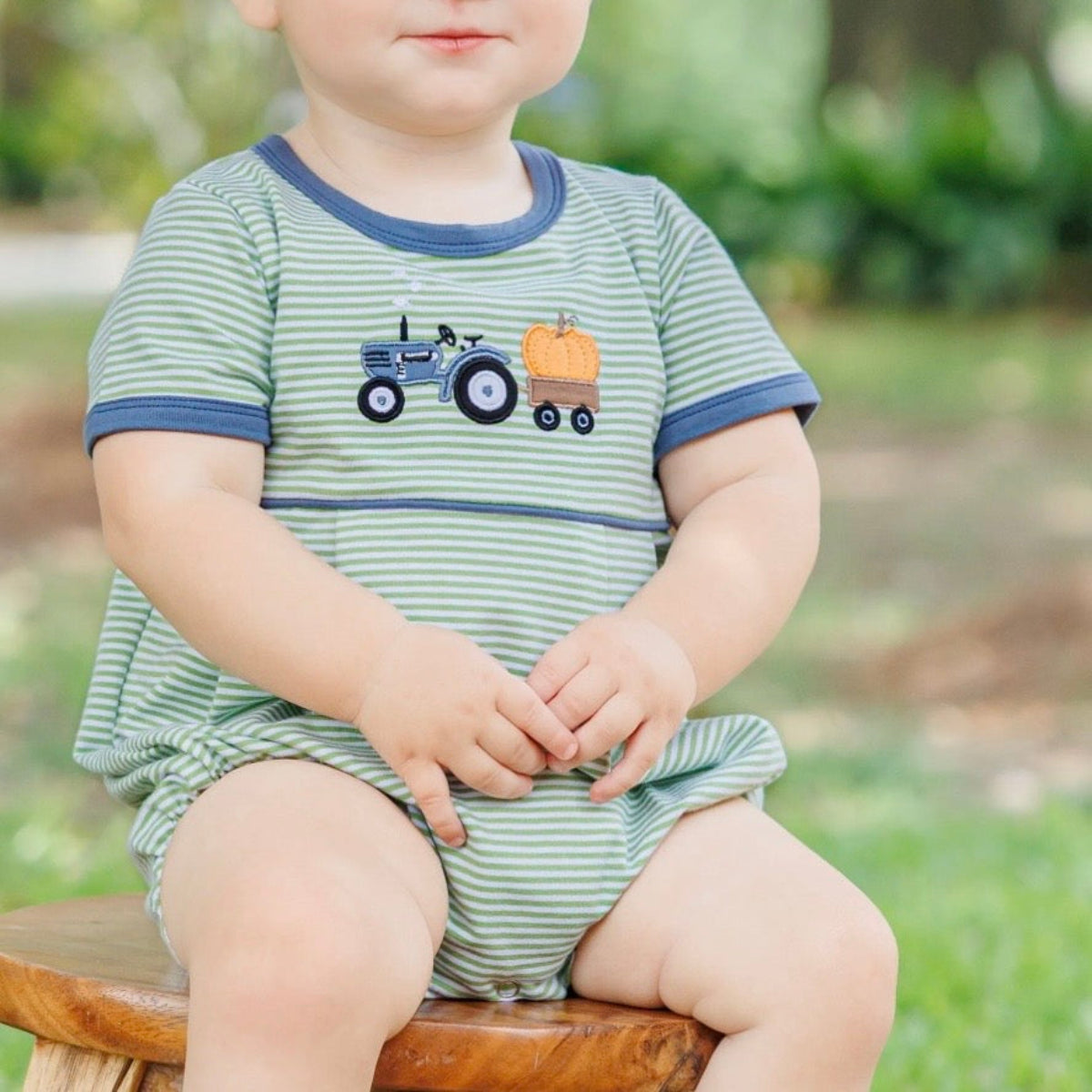 A baby boy wearing a fall bubble in green and white stripes, featuring a steel blue crew neck and cuffs and an appliqued tractor and orange pumpkin design.