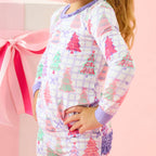 A young girl with blonde hair in a ponytail wearing a long-sleeve pajama set with a Christmas tree pattern, standing next to a pink gift box with a large pink ribbon.