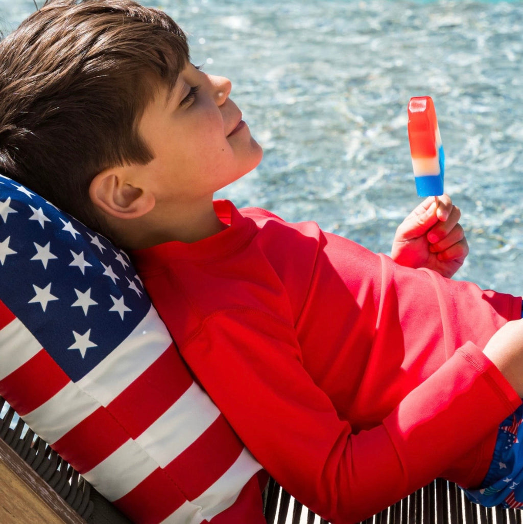 Child wearing a red long sleeve rashguard sun shirt holding a popsicle against an American flag pillow.