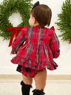 A toddler viewed from behind wearing a red plaid long-sleeve romper with ruffled trim and matching plaid bloomers, standing indoors against a white wall with green wreaths and red bows in the background.