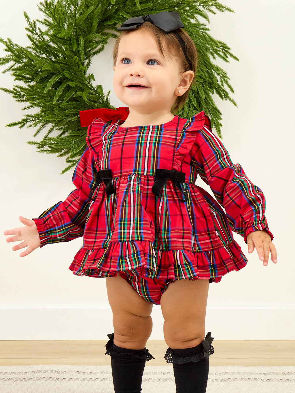 A baby girl wearing a festive red plaid long-sleeve romper dress with black bow accents, black knee socks, and a black bow headband, standing in front of a green wreath.